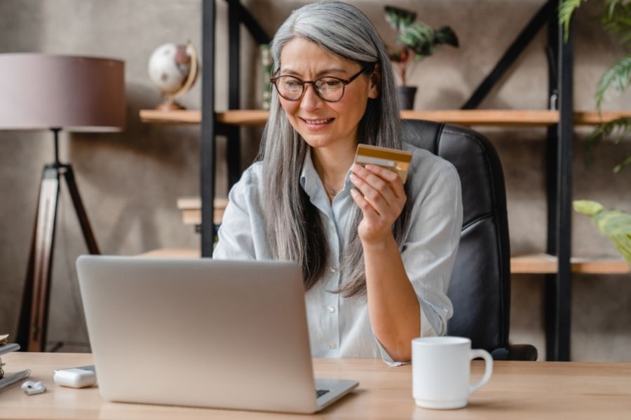 Grey haired woman sits in front of a laptop holding credit card. She is looking at the screen and smiling.