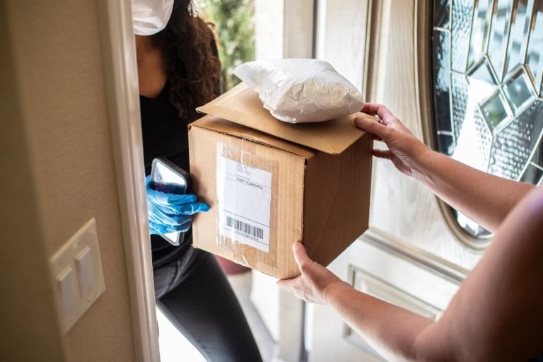 Photo of a home doorway where one person is receiving some parcels from a delivery person who is wearing a mask.