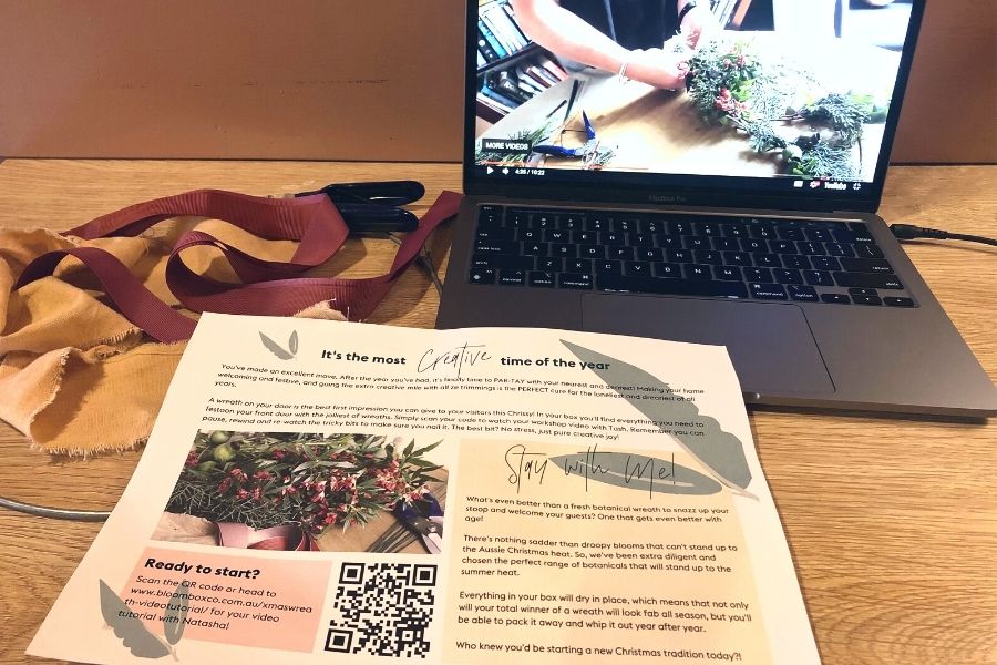Piece of A4 paper containing instructions and a QR code on how to create a floral Christmas wreath. A laptop is behind the paper showing the video that is linked to the QR code.