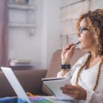 Photo of a woman sitting on a couch with a laptop on her lap. She is looking away from the camera biting a pen thoughfully