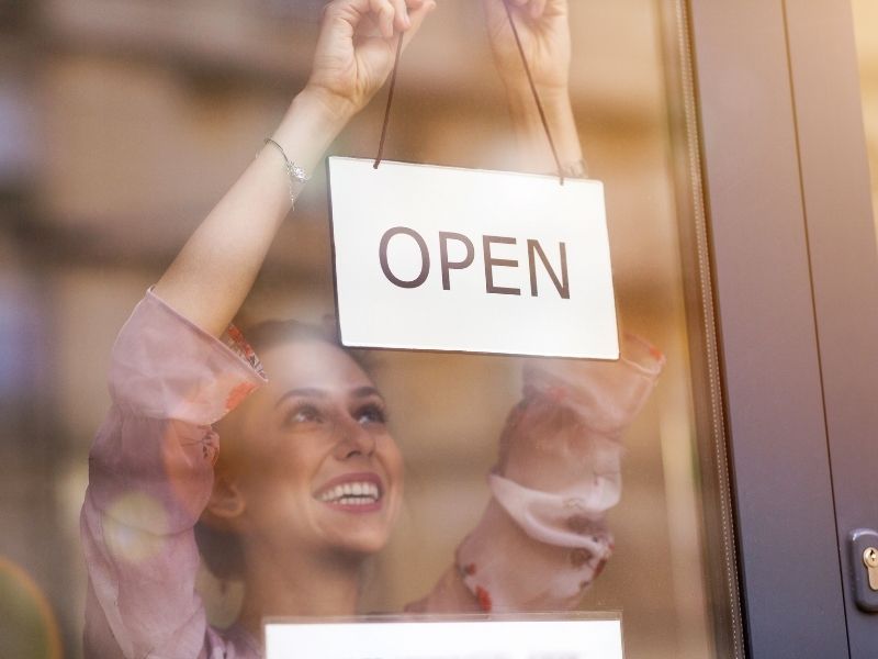 Photo of a woman hanging an open sign the a window of a small business